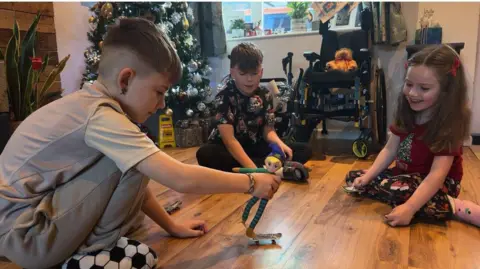 Chris McHugh/BBC Two young boys and a little girl play on a wooden living room floor with a cuddly toy and a miniature skateboard. A Christmas tree can be seen in the background of their family home. 