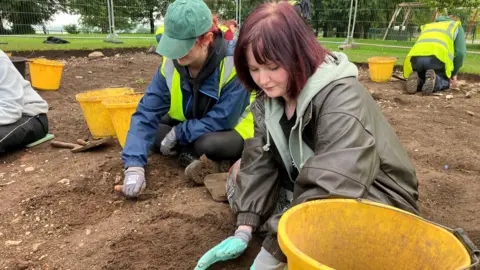 Aoife Lynch is removing soil into a bucket. She is wearing a green hoodie and a green coat, she has red hair. Behind her are numerous people all removing soil from the ground. There are multiple yellow buckets in the background. We can see the bottom of trees and a silver fence.