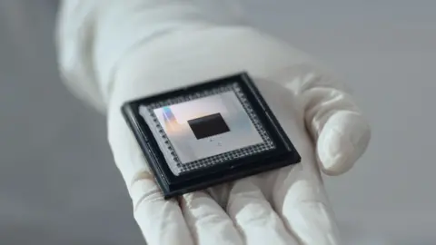 Google Google's Willow chip, which resembles three concentric black and silver squares the largest about the size of a beer mat, is held by a white gloved hand.