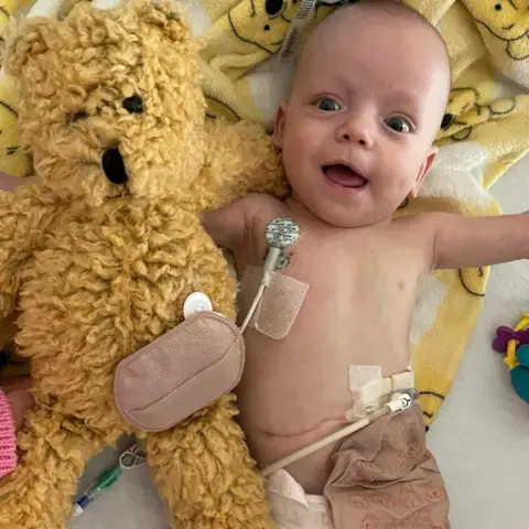 Bradford household  Baby Zachary successful  a infirmary  cot with monitors attached and lying beside a large  yellowish  teddy bear