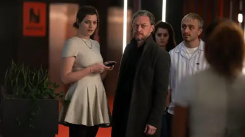 PA Media James McAvoy wearing a charcoal jacket with an earpiece in. There is a woman wearing a white dress stood to his left.