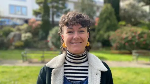 Tina Bury is a woman with dark curly hair and a nose piercing. She smiles at the camera. She has bright yellow hoop earring and a striped jumper with a cord jacket. Behind her is a park bench with grass.