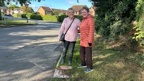 Two women standing next to a concrete plinth where a post box was sited before it was stolen, with a road to their right and trees to their left