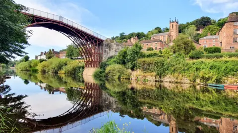 A small town with red brick buildings, a church and a metal framed bridge reflected in a still river