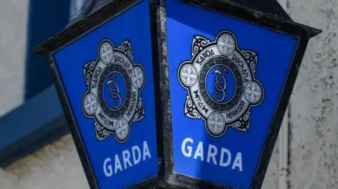 A blue Gardai sign with a grey and black Garda logo. There is a brick wall with a blue windowsill behind.