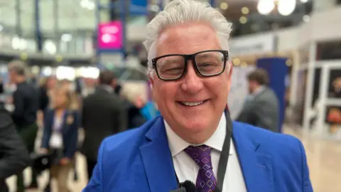 A man wearing glasses and a bright blue suit smiles at the camera