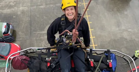 Person in fire rescue equipment and a stretcher smiling while hanging off a rope