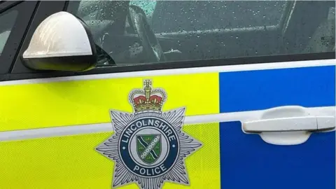 The side of a Lincolnshire Police patrol car. It has blue and yellow fluorescent markings and the force's badge on the door