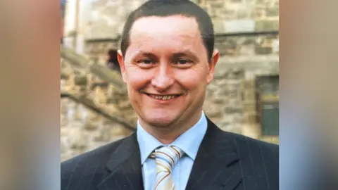 John Kelly A younger John Kelly wearing a suit with a blue shirt and multi-coloured stripey tie. He has dark hair and is smiling at the camera