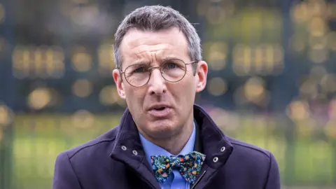 PA Media Andrew Muir is standing outside on front of large black gates. He wears glasses and is wearing a navy coat with a blue shirt and a bow tie.