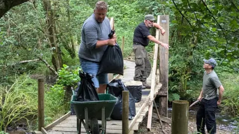 Root Cause Project Three men are working in the woods. Two stand on a raised wooden walkway, one of which is lifting a full black bin bag into a wheelbarrow. The other is talking to a man standing below him on the bank of a small stream.