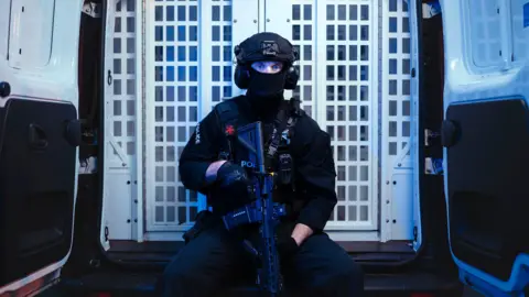 BBC/Expectation Entertainment/Drew Forsyth A police officer is holding a gun, sat down on the back of a van - the van doors are open and behind him you can see a white cage door. The officer is wearing a face covering, with only his eyes visible.