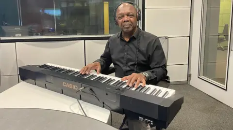 A bald man is wearing a black shirt. He is sitting behind a keyboard in a studio. 