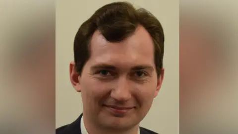 Harry Richardson smiles at the camera. He has brown hair and is wearing a dark coloured suit jacket with a white shirt underneath.