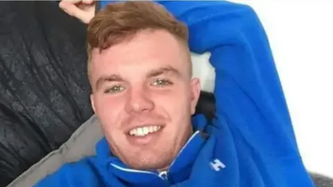 Greater Manchester Police Jack O'Brien, wearing a royal blue fleece long-sleeved top, smiles at the camera as he relaxes on a sofa.