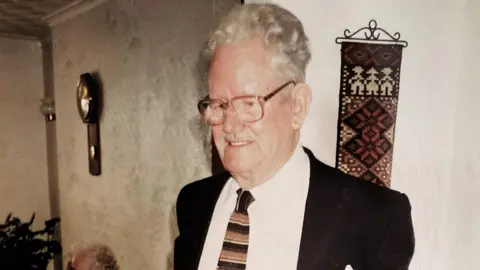 Steven Howells Bob Howells, who is wearing a white shirt, striped tie and black blazer, is standing in a living room with a clock and a tapestry on the wall behind him. 