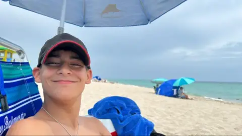 A child sat on a beach wearing a baseball cap. They are smiling at the camera.