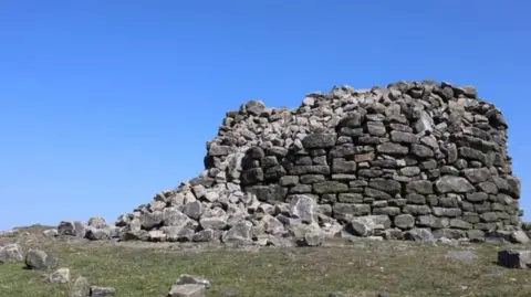Woodland Trust Large hut-size cairn made of large stones which has partially fallen. It stands at the top of a hill. The image is taken on a sunny day with blue skies.