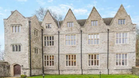 Fine and Country Outside of a detached Tudor mansion made of sand coloured stone bricks. There is grass in front of the property and black outdoor lights leading to brown double doors with black handles. There are 16 sets of windows from the front of the property.