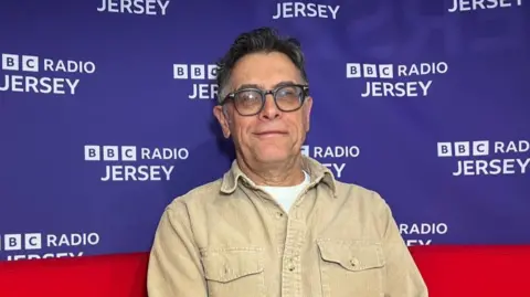 Tim Ashton Barnett is pictured on a BBC red sofa. A purple background is visible behind him with the BBC logo on it. He is wearing a beige corduroy shirt, dark glasses and has dark short hair.