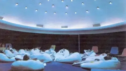 MANX NATIONAL HERITAGE People lie down on large cushions in a room with blue light, they lie as if they are tanning.