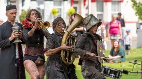 James Bass Photography A troupe of circus artists playing brass and drums. They are wearing dark clothing and have dirty arms, legs and faces. The drummer has a bucket on their head. The fourth person in the line up is holding a casket of ashes. People are sat behind them, watching, with a terrace of guest houses behind them.