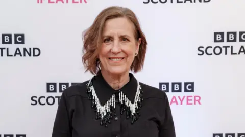 Two Rivers Media/BBC Scotland Presenter Kirsty Wark in a stylish black shirt with a long beaded black and white collar poses in front of a BBC Scotland backdrop which is white with black and pink logos