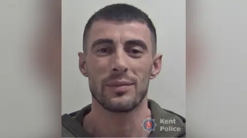 A police mugshot of a smirking man with short brown hair and stubble. 
