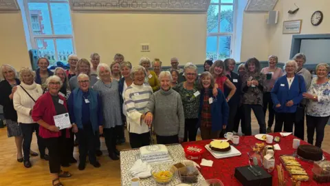 One Acchord A group of around 30 women standing behind a table with cakes inside a village hall