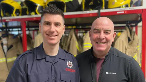 Two men are photographed together in a fire station's locker room. In the background there are beige fire fighter overalls hanging up and stacked together above, there are five yellow fire helmets. The men are smiling. One wears a Northern Ireland Fire Service uniform while the other is wearing a non-uniform black zip-up top.