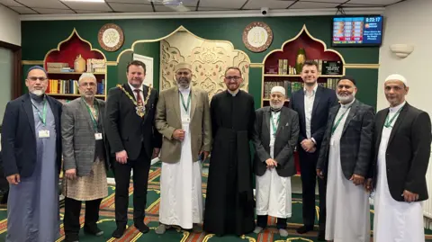 Jon Wright/BBC Nine men stand in a row inside a mosque, some are wearing Muslim dress, one is a Christian vicar, another in a suit and a third with mayoral chains.
