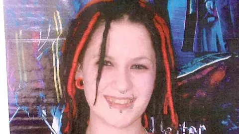 Sophie Lancaster, who was murdered in 2007, is pictured with red and black hair and a red plug earring, a nose and lip piercings. She is standing in front of poster.