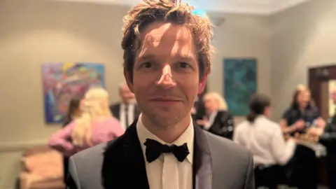 Damien Molony dressed in a formal black tuxedo with a white dress shirt and a black bow tie stands indoors at what appears to be a social or formal event. The background is softly lit and out of focus, showing several people mingling, artwork on the walls, and glasses set out on a table. 