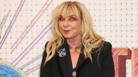 Getty Images Helen is stood in front of a colourful backdrop. She has long blonde hair and is wearing a black suit jacket and smiling at the camera.