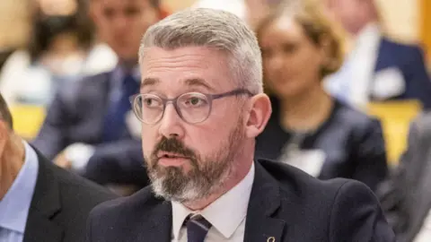 PA The former senator Niall Ó Donnghaile speaking at a committee meeting surrounded by other politicians. He is wearing a dark suit and tie with a white shirt.