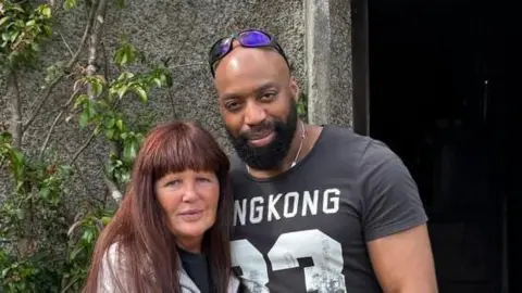 Denise, with red hair and a light blue jacket, with her arms around her partner Sylvester Abayomi, who is wearing a black T-Shirt with white numbers and with sunglasses on his head, in a photo released by the family.