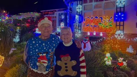 Bill and Barbara Wright Bill and Barbara wearing Christmas jumpers, smiling outside their house which is covered in Christmas lights and illuminated objects including snowmen and Santa dogs.