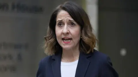 PA Media A woman with dark hair speaks while walking along a street. She is wearing a dark blue jacket over a white top. 