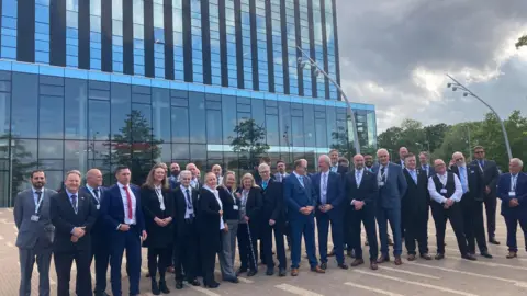 Laura Coffey/BBC Men and women wearing suits, standing on the plaza outside the Corby Cube. Many of them are wearing council lanyards.