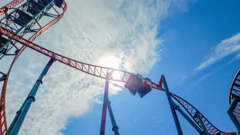 Diario de una montaña rusa Una montaña rusa que se muestra desde abajo. Puedes ver un carrito al revés que se abre paso por una pista giratoria. La montaña rusa tiene una pista de naranja con vigas de soporte azul. Detrás hay un cielo azul con el sol brillando a través de una nube. 