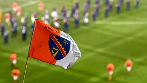 An Armagh flag - which is half orange and half white with a blue crest on it - waves in front of a blurred background of a green pitch. On the pitch are players in orange Armagh GAA jerseys as well as a marching band in blue uniforms