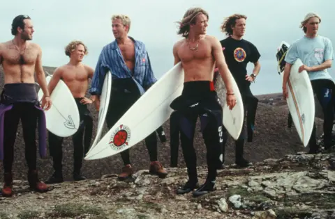 Carl Prechezer/FilmFour Six Cornish surfers in a line, carrying surfboards. Three have the top half of their wetsuits rolled down, and the others have shirts on. 