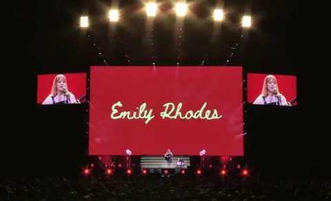 Emily Rhodes One main screen has a dark red background with Emily Rhodes written on it in a beige-coloured, joined up front. Two smaller screens sit either side of it showing Emily Rhodes in front of a microphone. Lights are overhead, and there's a small figure of someone performing on stage and holding a guitar.