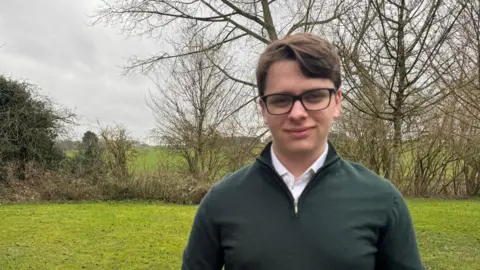 Bobby Seymour Councillor Bobby Seymour stands in a field while wearing a dark green pullover, a white shirt and glasses. He has short brown hair parted on one side. Behind him is a low hedgerow with some trees