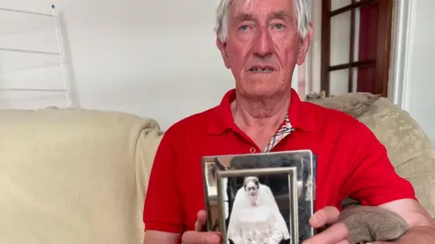 Raymond Turnbull in a red T-shirt and wearing a watch. He is holding a silver frame with a black and white photo of his wife Jean in a wedding dress