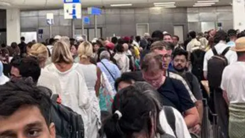 People queue at Athens airport in long lines in a white terminal building.