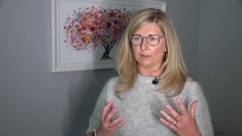 Miriam Wilkinson speaks during an interview, wearing a light grey top, with a framed artwork featuring a tree made of colourful heart shapes on the wall behind.