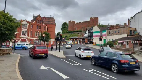 General view of Shrewsbury station gyratory -  a number of cars on the road with a large historic red brick building and castle in the background of the photo