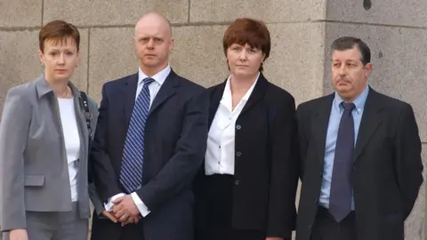 Medios de comunicación de PA: Cuatro personas de pie, hombro con hombro. A la izquierda, una mujer pelirroja con un traje gris, junto a ella, un hombre con traje azul y corbata a rayas azules, junto a él, una mujer con un traje oscuro y junto a ella, un hombre con traje oscuro y corbata oscura.