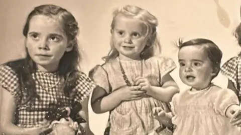 Old yellowing black and white photo of the three sisters as young children. The eldest on the left is wearing a checked dress and has long brown hair. The middle girl is younger and has blond hair and is wearing a plain dress while holding a necklace around her neck. The youngest on the right is a toddler with short dark hair.
